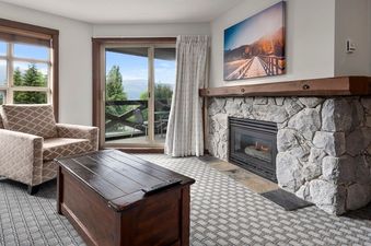 A cozy living room at Blackcomb Springs Two Bedroom Slopeside Suite features a stone fireplace, a comfortable armchair, and a dark wood coffee table, with a large window and glass door offering mountain and forest views.