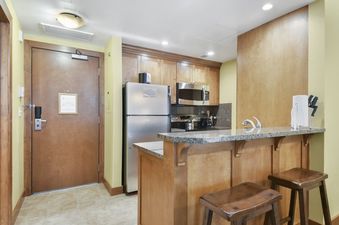 The Sundial Lodge Studio features a full kitchen with stainless steel appliances, wood cabinetry, granite countertops, and a breakfast bar with two stools, adjacent to a secure wooden entrance door.
