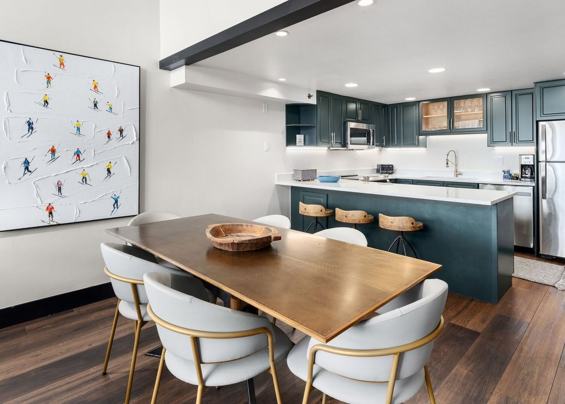 A modern dining area with a large wooden table and chairs, adjacent to an open-plan kitchen with dark green cabinetry and a white countertop.