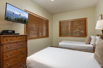 A secondary bedroom with two twin beds, a wall-mounted flat-screen TV, and wooden blinds covering the windows.