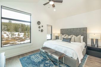 A spacious bedroom at Alpenglow Retreat features a large bed, a wooden headboard, and a large window overlooking a snow-covered forest.