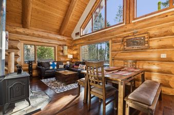 A rustic living and dining area in Hellroaring Hut features log walls, a vaulted ceiling, a wooden dining table, and a cozy seating area with a leather sofa and wood-burning stove.