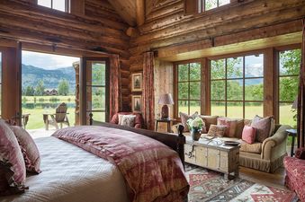A luxurious bedroom at Grizzly Wulff Lodge with a large bed, a comfortable seating area, and expansive views of a lake, mountains, and greenery through large windows and an open door.