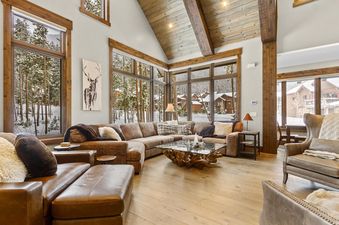 A spacious living room at Timberline Lodge with a large brown sectional sofa, glass coffee table with a rustic wood base, and floor-to-ceiling windows offering a snowy forest view.