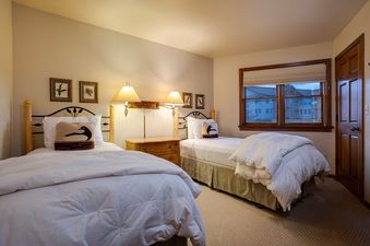 A cozy twin bedroom at Chateau Chamonix CX123 features two beds with white bedding, bird-motif pillows, a rustic nightstand with wall lamps, framed art, a window with an exterior view, and a wooden door.
