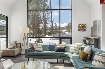The living area of the Modern Alpine Escape features a blue sectional sofa, a glass coffee table, and large windows revealing a snowy mountain landscape.