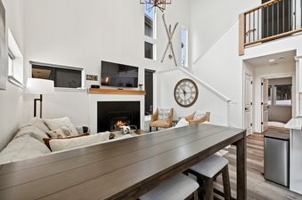 The bright and spacious living area of Quarry B23, featuring high ceilings, large windows, a prominent fireplace with a warm glow, comfortable seating, an elegant dark wood dining table, and unique decorative accents like wall-mounted skis and a large cir