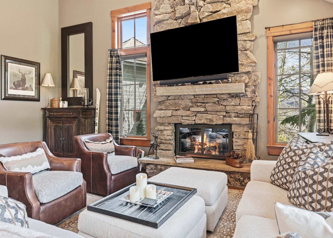 A cozy living room at Buckhorn Townhome 264 features a roaring stone fireplace, a flat-screen TV, comfortable leather armchairs, and large windows with plaid curtains overlooking a natural landscape.