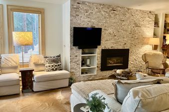 The inviting living area features plush seating, a fireplace set in a stone wall, and a large window offering an outdoor view.