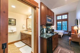 A hotel room with a kitchenette and a bathroom. The kitchenette features a microwave, a small refrigerator, and a sink. The bathroom has a double sink and a large mirror.