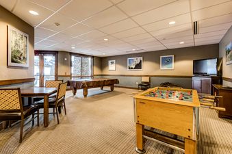 The games room at Trailhead Lodge 2105 showcases a pool table, a foosball table, and a dining area, set against a backdrop of framed art and large windows.
