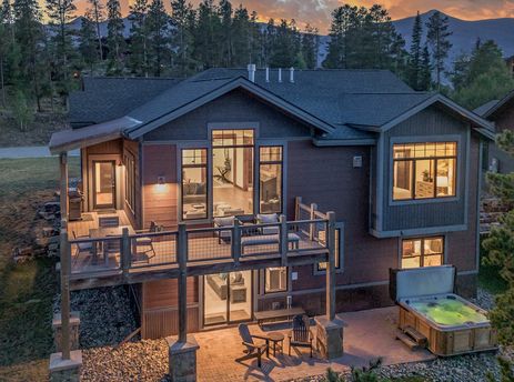 A multi-story chalet with a spacious deck and ground-level patio, featuring outdoor dining, a hot tub, and large windows, set against a backdrop of pine trees and mountains at sunset.