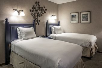A cozy bedroom at The Corporate Suite Beaver Run Resort with two twin beds, each featuring crisp white linens and a wall-mounted light, complemented by wall art and a decorative metal tree sculpture.