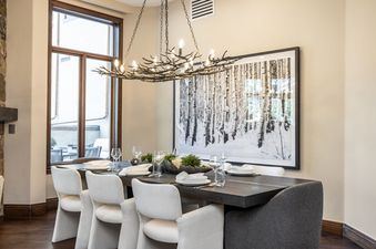 A luxurious dining room in the Sunstone Chalet at One Beaver Creek, featuring a long dark wood table, white upholstered chairs, a unique branch-like chandelier, and a large black-and-white artwork of a birch forest.