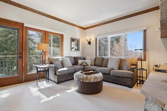 A cozy living room featuring a large sectional sofa, a round cowhide ottoman with a tray, and a wall of windows and glass doors opening to a balcony with mountain views.