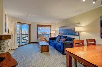 The living area of the Blackcomb Springs One Bedroom, featuring a blue sofa, an armchair, a coffee table, and a dining table with chairs, all set on a patterned carpet.