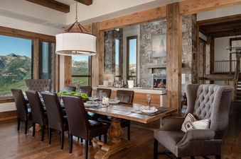 The dining room at 30 Spur Road Moonlight Basin features a large wooden table with leather chairs, a stone fireplace, and a stunning view of the mountains.