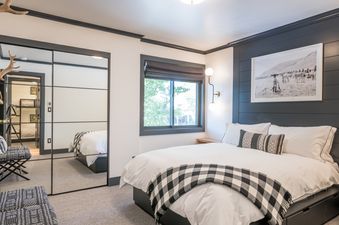 A bedroom with a king sized bed, white bedding, and a black and white checkered throw blanket. There is a large window with a view of trees, a mirrored closet, and a picture on the wall above the headboard.