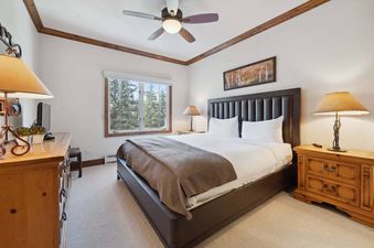 A cozy bedroom with a bed, two nightstands with lamps, a dresser, and a window overlooking pine trees.