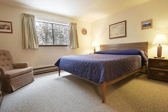 The bedroom at Beaver Village 1821 features a queen-size bed with a blue comforter, two nightstands with lamps, a comfortable armchair, and a large window offering a view of snow-covered trees.
