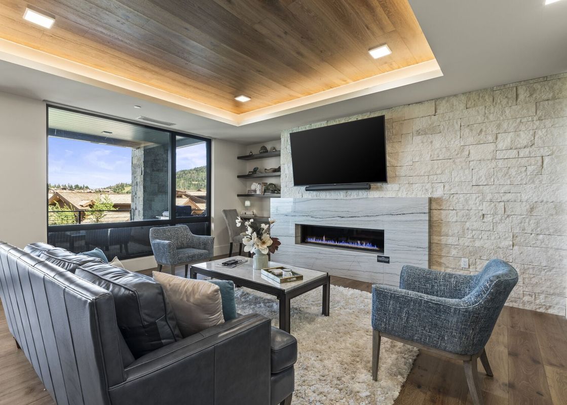 A modern living room with a black leather sofa, blue armchairs, a stone fireplace, and a large window offering mountain views.