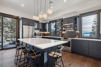A modern kitchen in Last Chair Lair with dark cabinets, stainless steel appliances, a white island with bar stools, and large windows offering a view of snow-covered mountains.