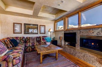 The living area of Peaks Penthouse 752 features a large, colorful sectional sofa, a rustic wooden coffee table, a fireplace, and large windows offering views of snow-capped mountains.