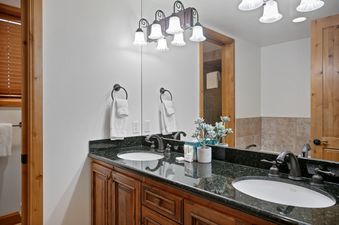 A double vanity in the Bellemont 5 bathroom features dark granite countertops, two white sinks with bronze faucets, and a large mirror reflecting warm wood trim.