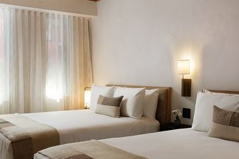 A bright and inviting bedroom in the Mollie Aspen Park View Corner Suite features two queen beds with neutral bedding, a textured wood ceiling, and sheer curtains covering a large window.