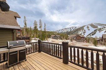 A Weber BBQ grill sits on the spacious wooden deck of Mountain Majesty Winter Park, offering stunning views of snow-capped mountains and pine forests under a cloudy sky.