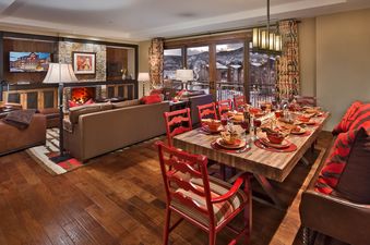 The dining and living area of Emerald Mountain One Steamboat Place 702 features a rustic wooden dining table, a cozy fireplace, comfortable seating, and large windows revealing a snowy mountain landscape.