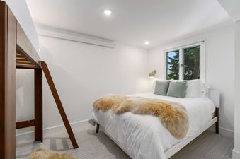 A cozy bedroom in the Solstice Suite featuring a queen bed with a fluffy throw, a bunk bed ladder, and a window showing green trees.