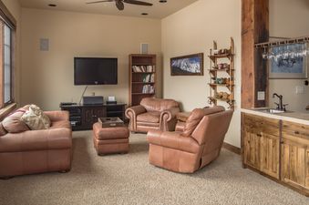 A cozy entertainment area with brown leather seating, a flat-screen TV, a bookcase, and a wet bar.