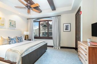 The bedroom in Lumiere One Bedroom Standard Residence features a king-size bed with a plush white duvet, cozy throw blankets, and blue patterned pillows. The room is spacious with a large window offering views of the surrounding snowy mountains.