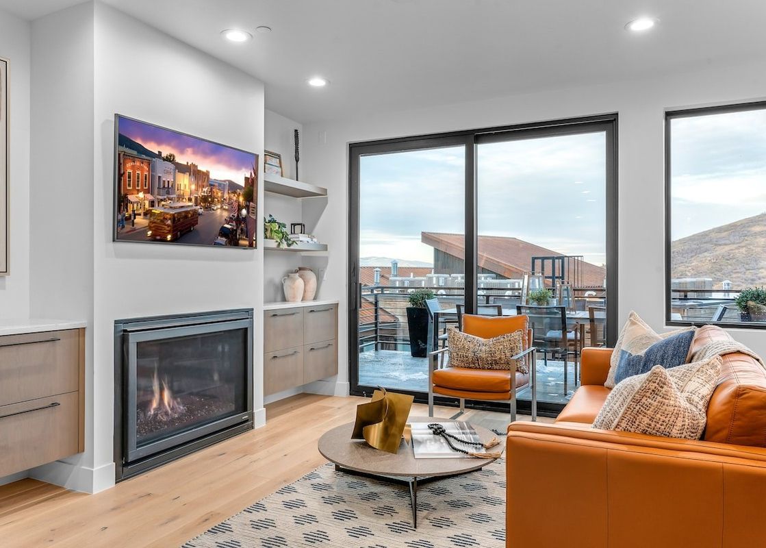 A living room with a modern fireplace, a couch, and a chair, looking out onto a balcony with views of a nearby mountain.