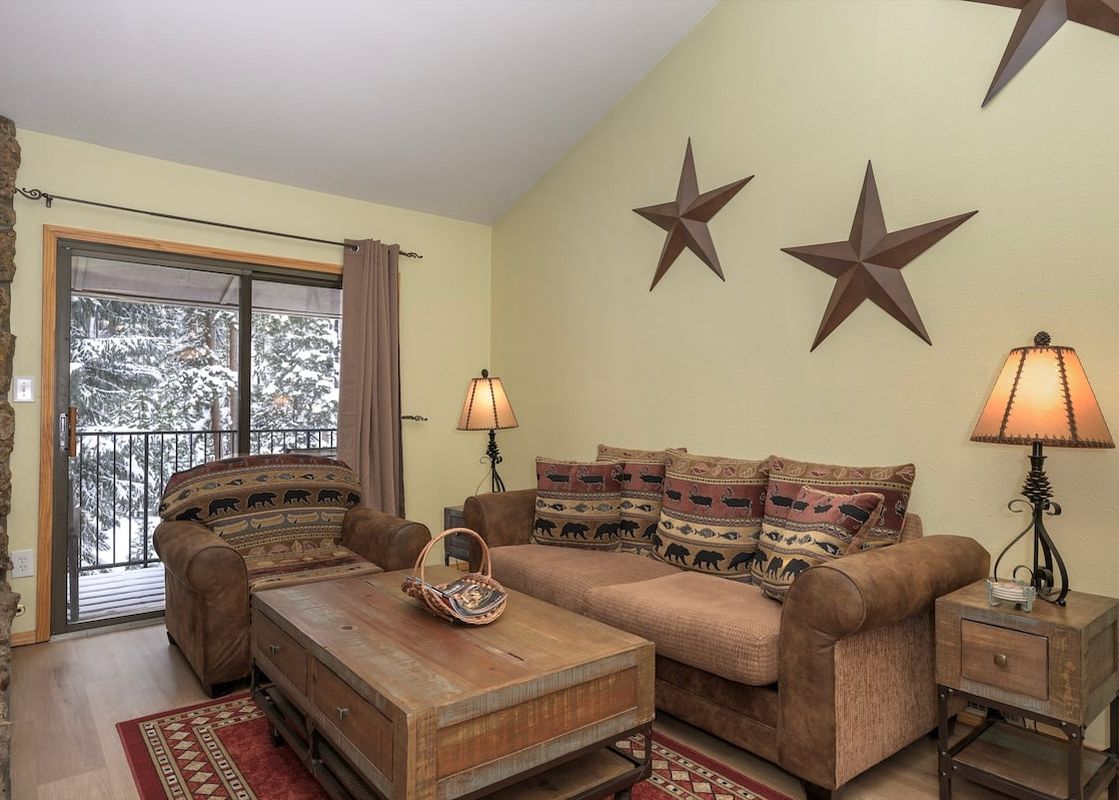 A cozy living room at Beaver Village 232 features a comfortable brown sofa and armchair, a rustic wooden coffee table, and large windows revealing a snowy forest.