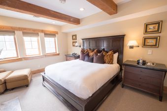 The primary bedroom features a large, dark wooden bed with white linens and an assortment of decorative pillows, set against a light brown wall with two framed pictures.