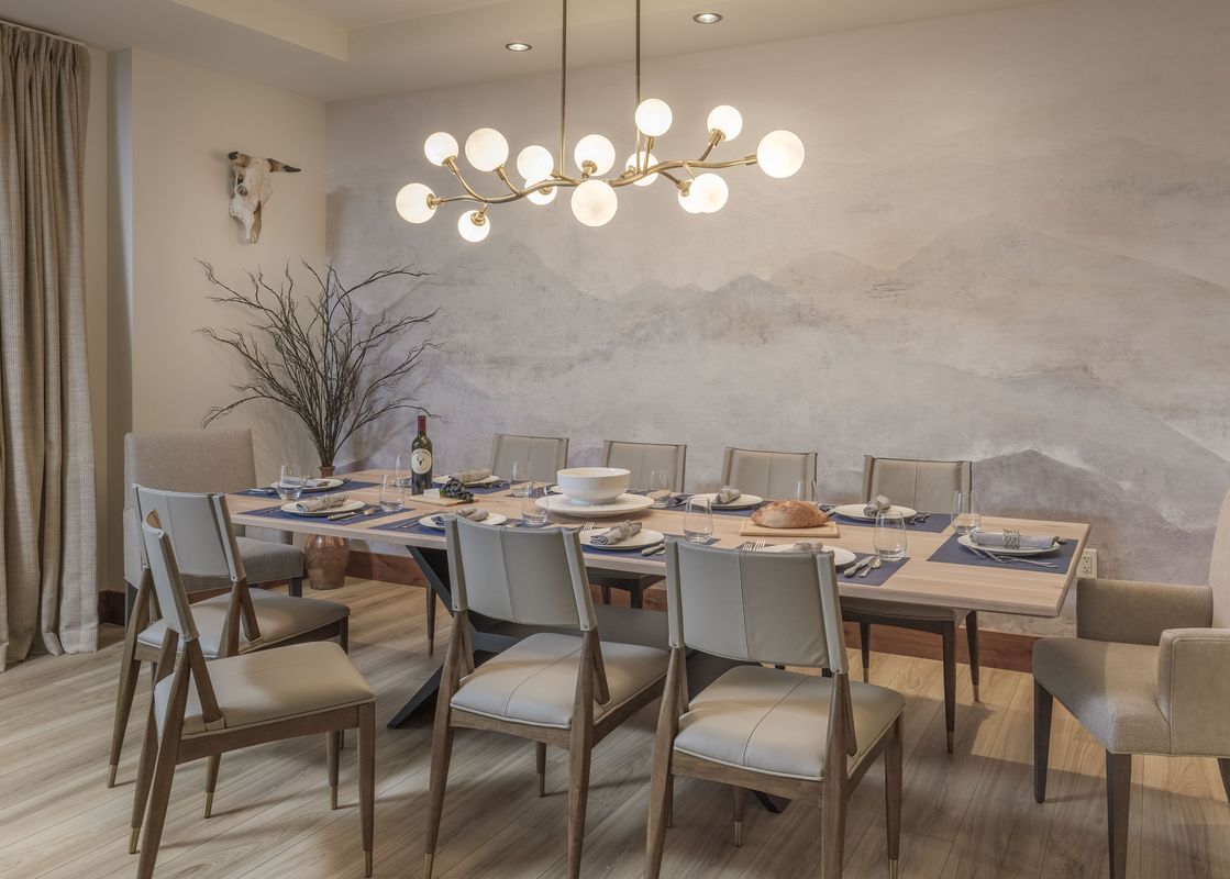A dining room table is set for a meal with plates, glasses, and silverware. The room is decorated with a faux antler and a mountain mural behind the table.