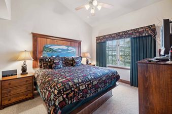 A cozy bedroom with a patterned quilt, wooden headboard, and two nightstands with lamps, featuring a window with curtains and an outdoor view.