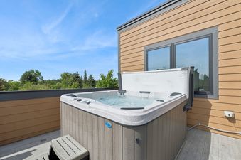 An outdoor hot tub with bubbling water on a deck, surrounded by building siding, a large window reflecting the sky, and lush green trees in the background, offering a serene view at Alpenglow Retreat Whitefish.