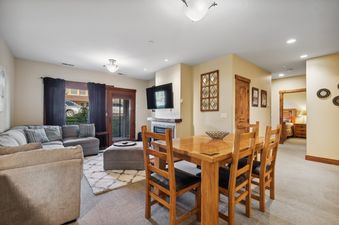 An inviting open-concept living and dining area in Pines Unit 240 features a large sectional sofa, a wooden dining table with seating for four, a flat-screen TV above a fireplace, and a glass door leading to a balcony.