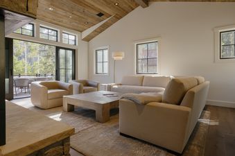 A bright and modern living room with light-colored sofas and armchairs, a large wooden coffee table, and a glass door leading to an outdoor deck surrounded by trees.