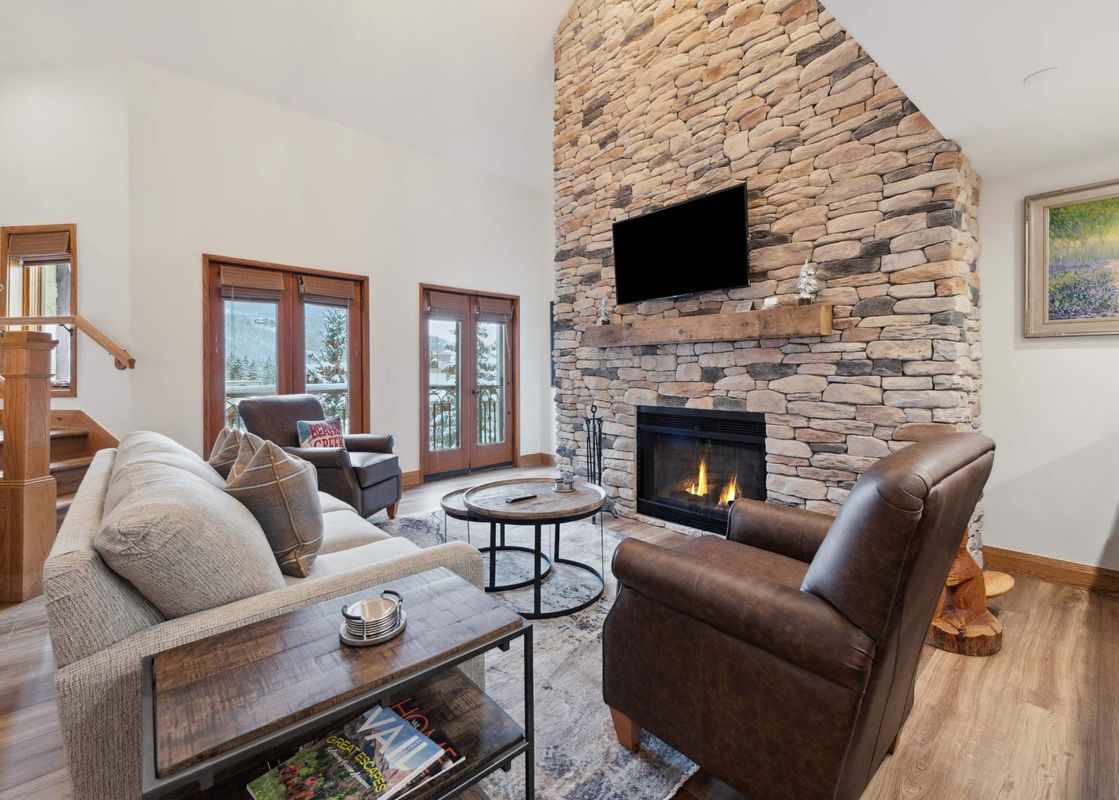 A spacious and elegantly furnished living room in The Charter L223, featuring a grand stone fireplace with a flickering fire, comfortable seating, and large glass-paned doors offering a view of a snowy landscape.