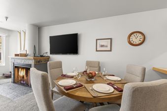 A dining room with a round wooden table set with plates, glasses, and maroon napkins. To the left is a fireplace with a roaring fire, and above it, a flat-screen TV is mounted on the wall. A wooden clock hangs on the wall to the right of the TV.