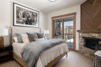 A cozy bedroom at Moonlight Basin 10 Wildwood, featuring a king-sized bed with plush bedding, a fireplace, and a stunning view of the snow-capped mountains through a large window.