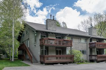 The Krystal Villa 5 building is a multi-story condominium with light green siding and dark wood balconies, surrounded by lush green trees under a bright blue sky.
