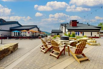 A sunny outdoor patio area features several wooden Adirondack chairs arranged around a central fire pit, with restaurants and shops in the background.