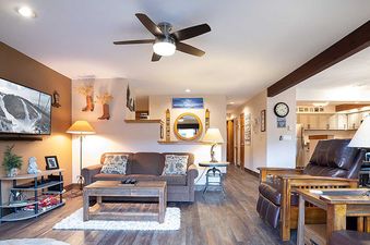 The living area features a brown sofa, a rustic wooden coffee table, and a large flat-screen television mounted on an accent wall, complemented by warm lighting and a ceiling fan.