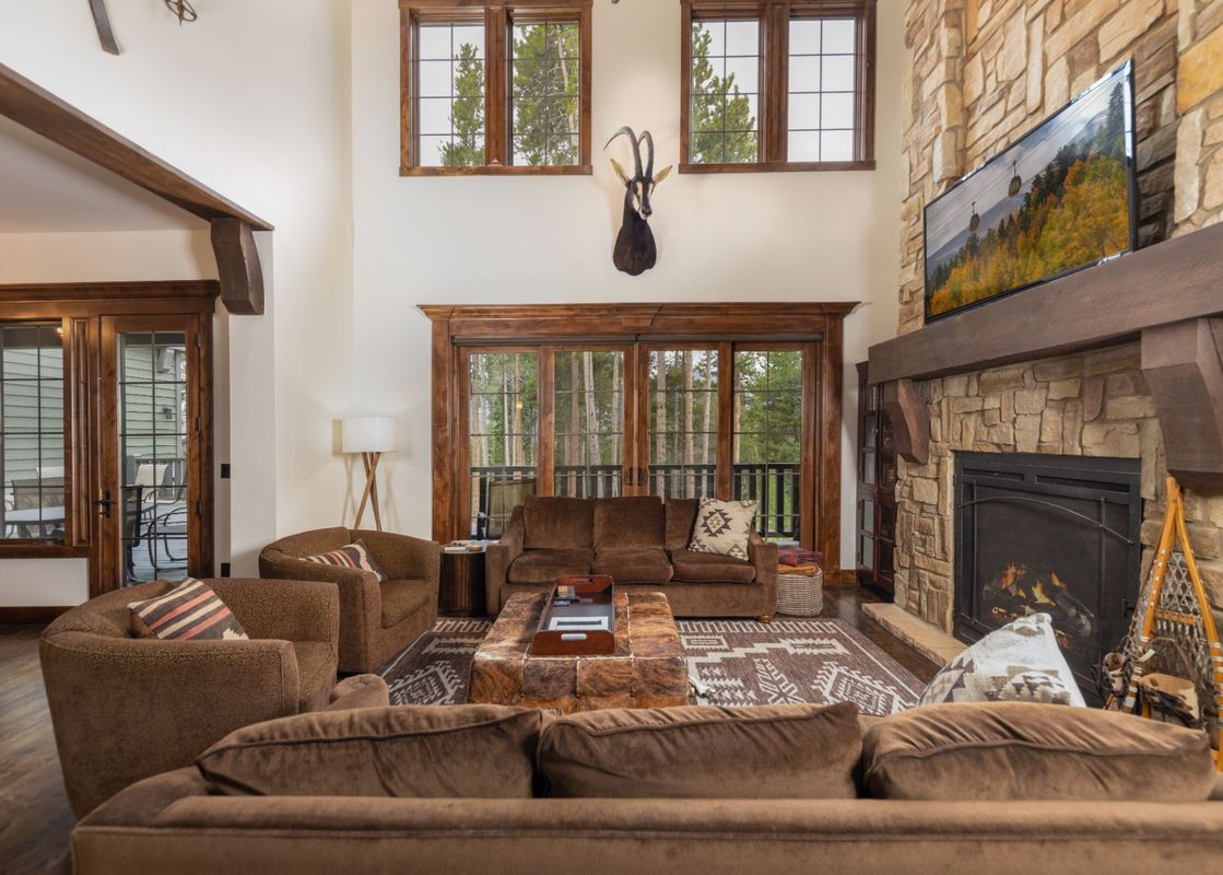 The living area at the Estate at Baldy Ridge features plush brown seating, a rustic stone fireplace, and large windows offering a serene view of pine trees.