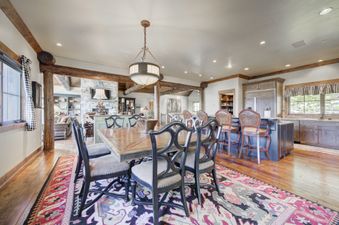A rustic yet elegant dining room at Peaks View Lodge with a large wooden table, dark chairs, a patterned rug, and a chandelier, leading to an open-concept living room and kitchen.